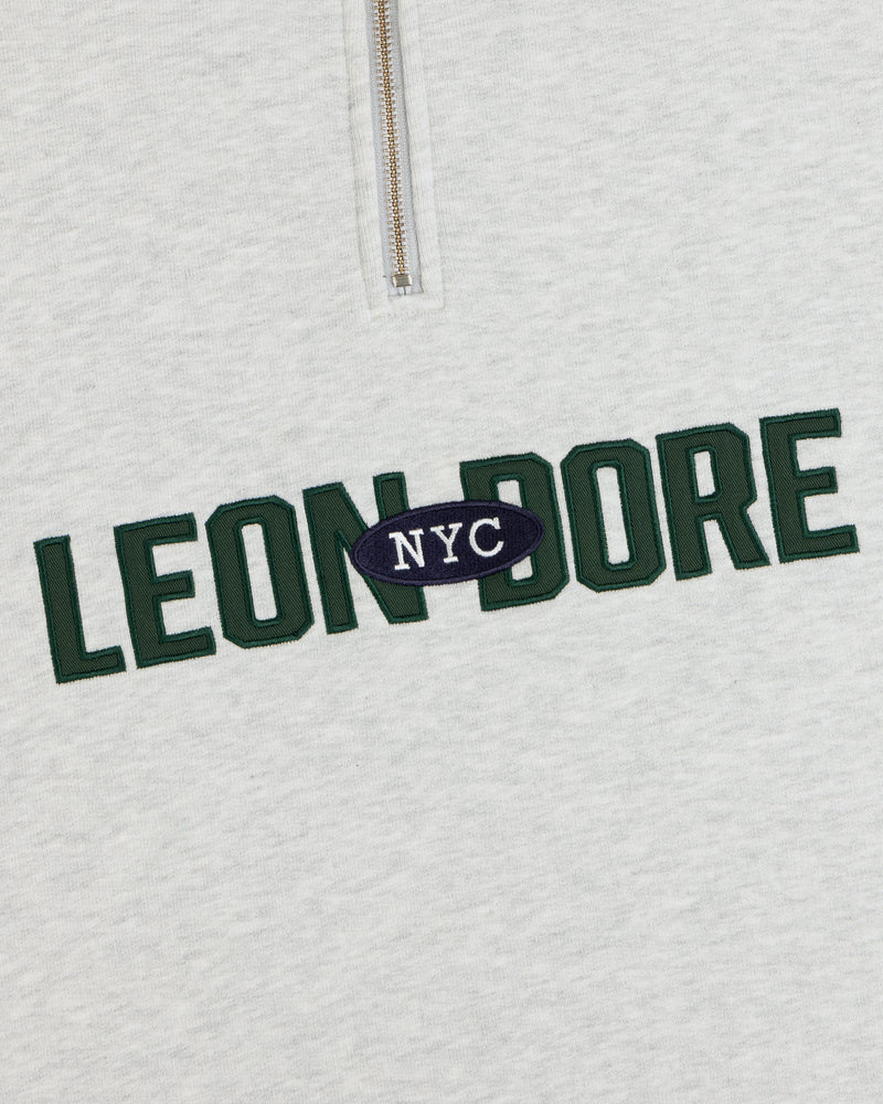 Close-up of Aimé Souvenir Quarter Zip’s light gray fabric, LEON DORE in green, NYC in white on a navy oval.