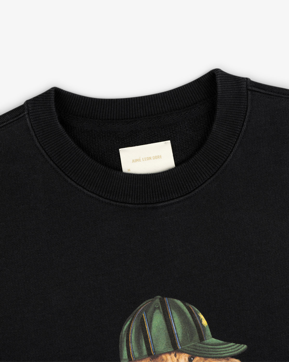 Close-up of the Buddy Crewneck Sweatshirt’s cream Aimé Leon Dore tag and a bear graphic in a striped hat.