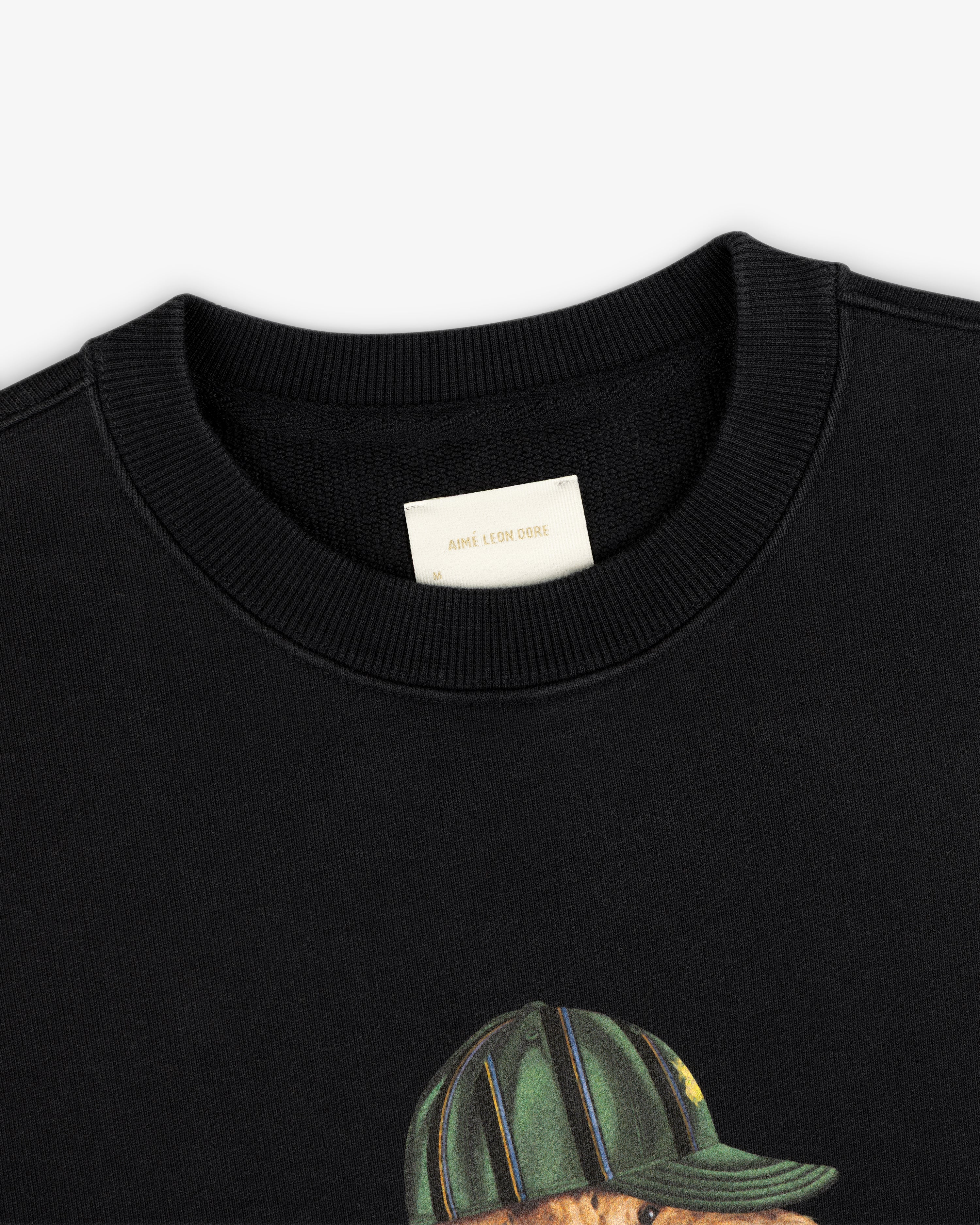 Close-up of the Buddy Crewneck Sweatshirt’s cream Aimé Leon Dore tag and a bear graphic in a striped hat.