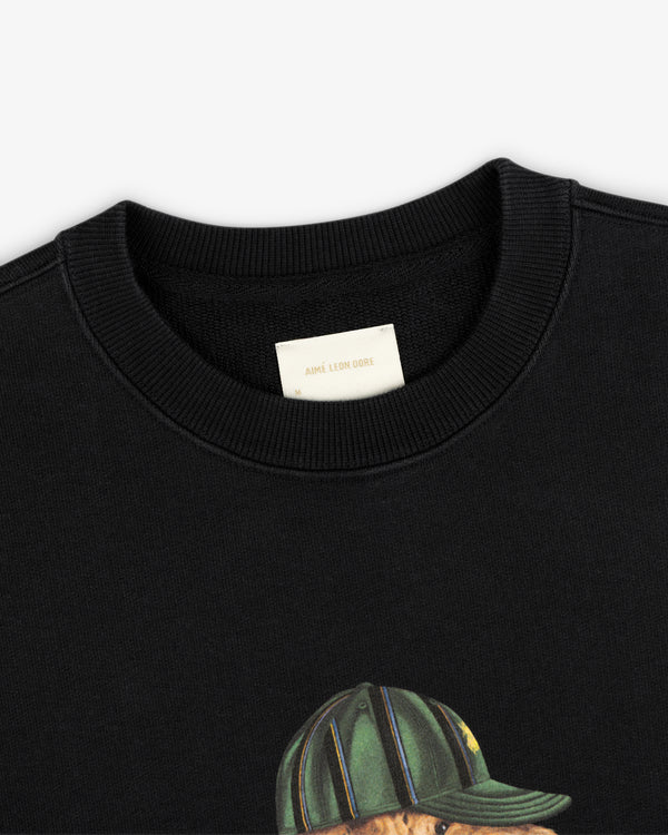 Close-up of the Buddy Crewneck Sweatshirt’s cream Aimé Leon Dore tag and a bear graphic in a striped hat.