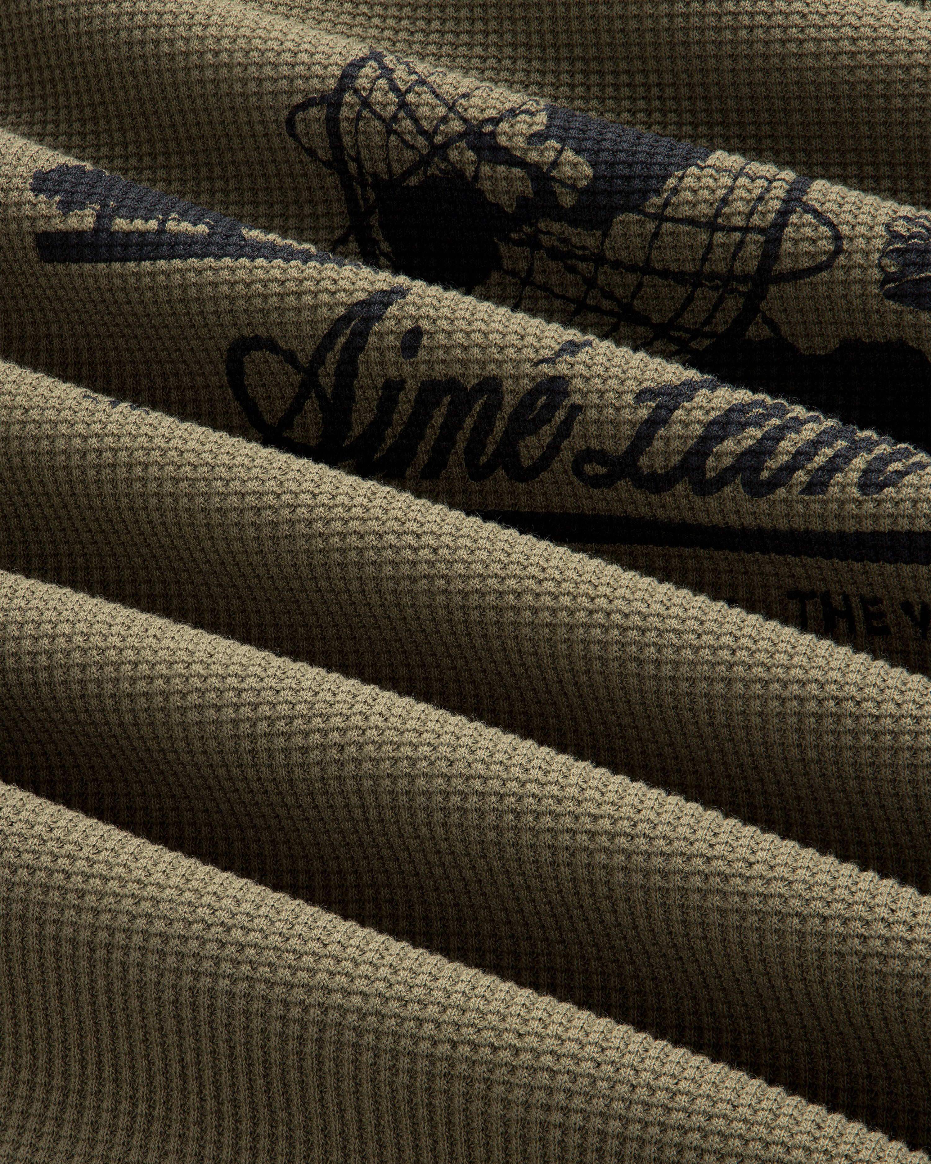 Close-up of olive green Long-Sleeve Unisphere Waffle Thermal with black printed text and graphic details.