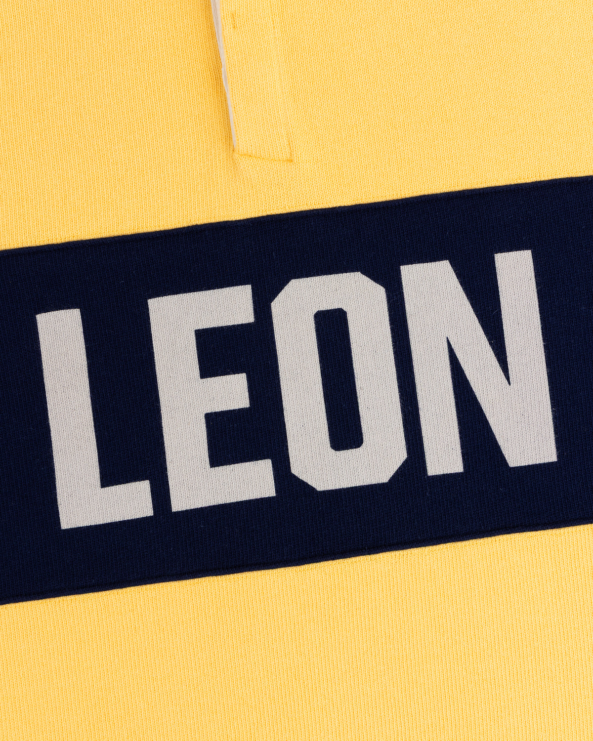 Close-up of the Leon Dore Collegiate Rugby: yellow shirt, navy stripe, bold white “LEON” text.