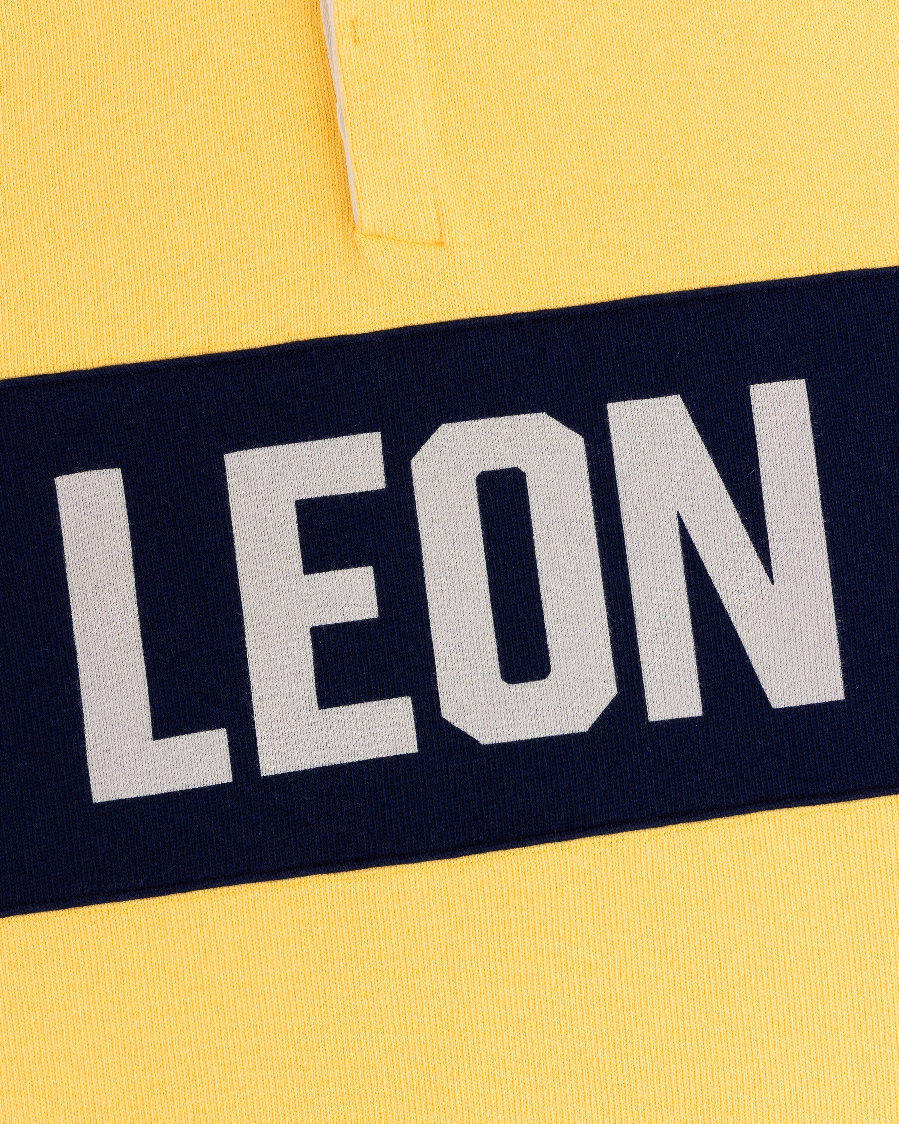 Close-up of the Leon Dore Collegiate Rugby: yellow shirt, navy stripe, bold white “LEON” text.
