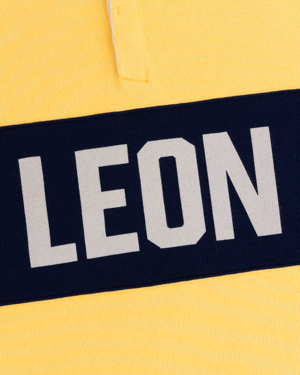 Close-up of the Leon Dore Collegiate Rugby: yellow shirt, navy stripe, bold white “LEON” text.
