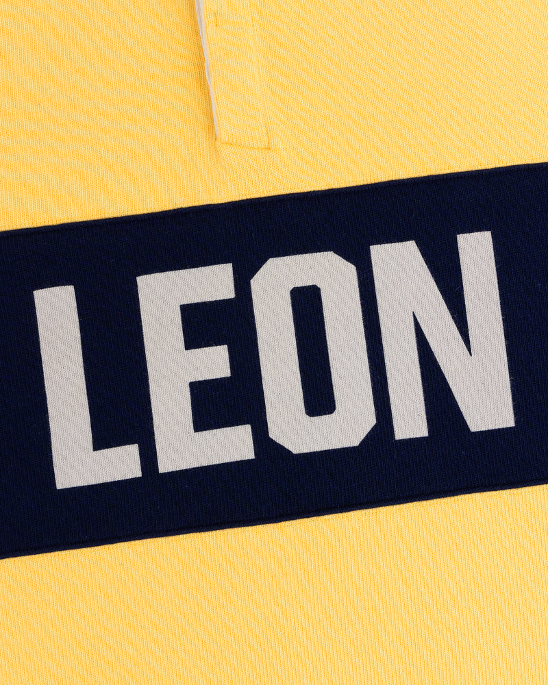 Close-up of the Leon Dore Collegiate Rugby: yellow shirt, navy stripe, bold white “LEON” text.