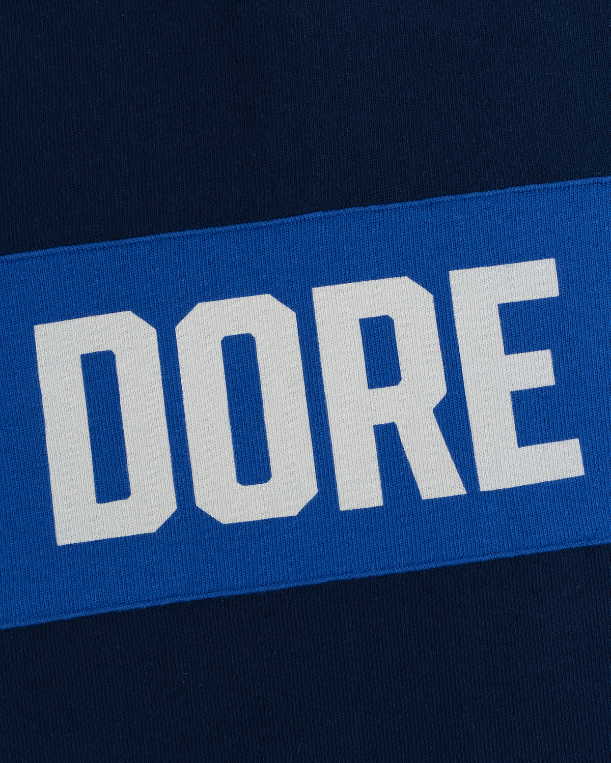 Leon Dore Collegiate Rugby: white “DORE” text on blue rectangle, set against dark blue fabric background.