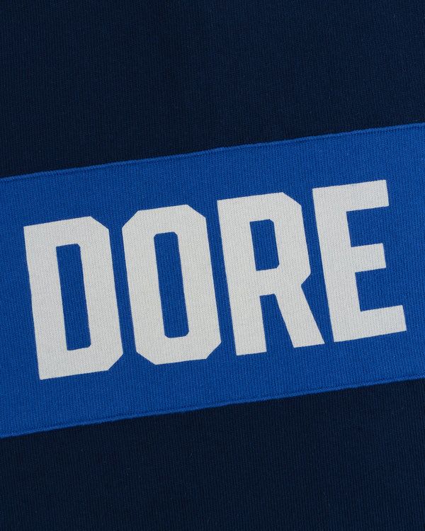 Leon Dore Collegiate Rugby: white “DORE” text on blue rectangle, set against dark blue fabric background.