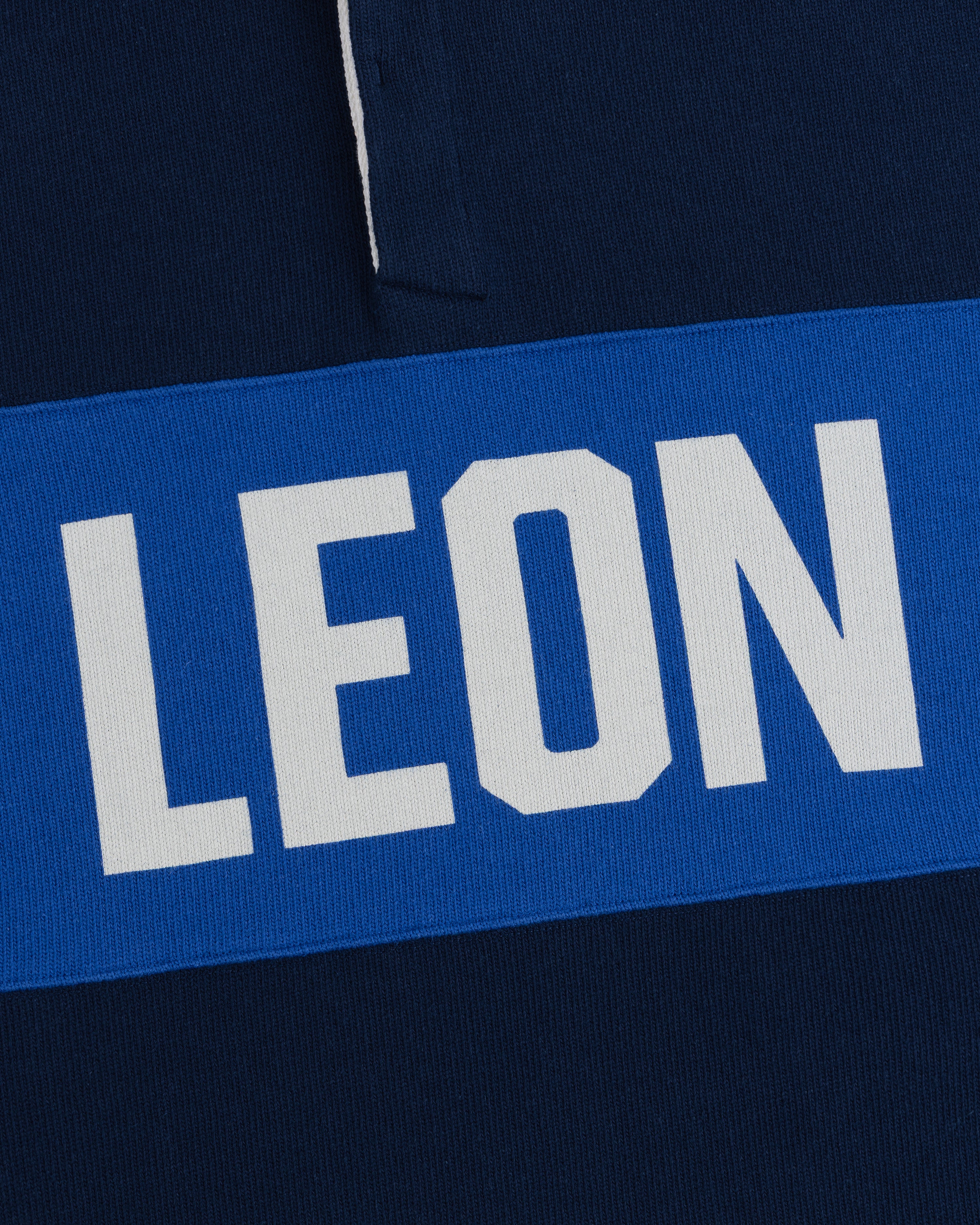 White “LEON” text on blue stripe over dark blue fabric, featured on the Leon Dore Collegiate Rugby.