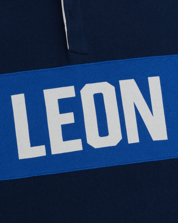 White “LEON” text on blue stripe over dark blue fabric, featured on the Leon Dore Collegiate Rugby.
