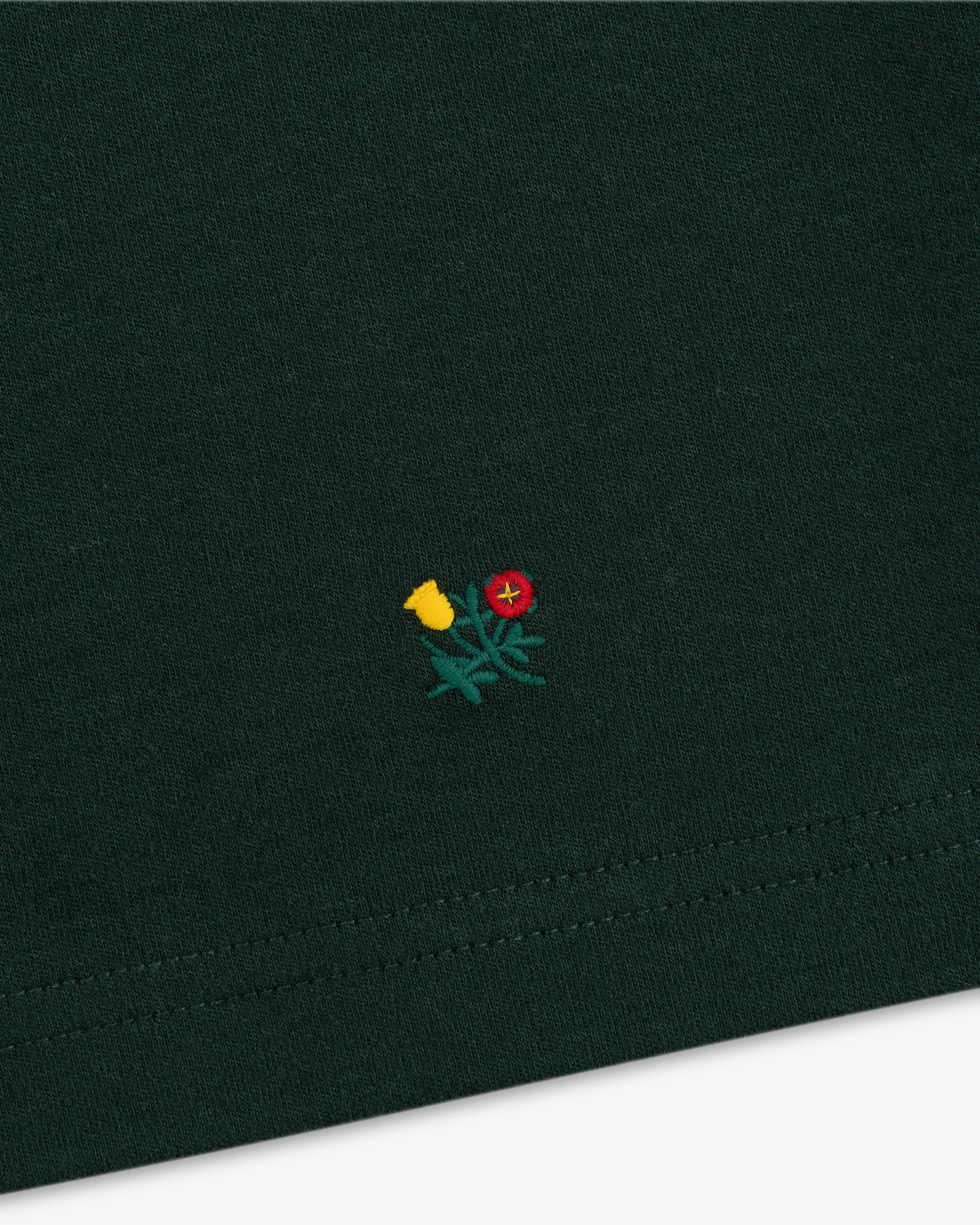 Heavyweight Crest Mockneck Tee in dark green fabric with a small embroidered emblem of a red and yellow flower.