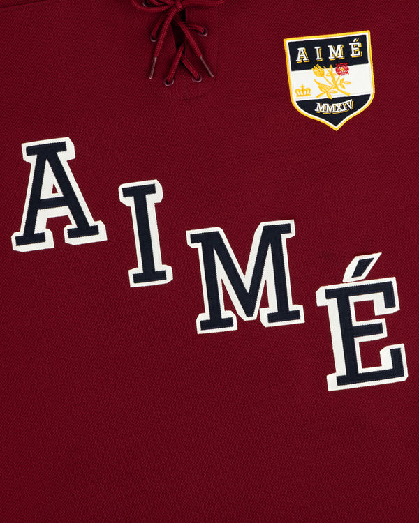 Team Aimé Hockey Jersey: maroon with a lace-up collar, varsity-style AIMÉ lettering, and a crown and floral crest.