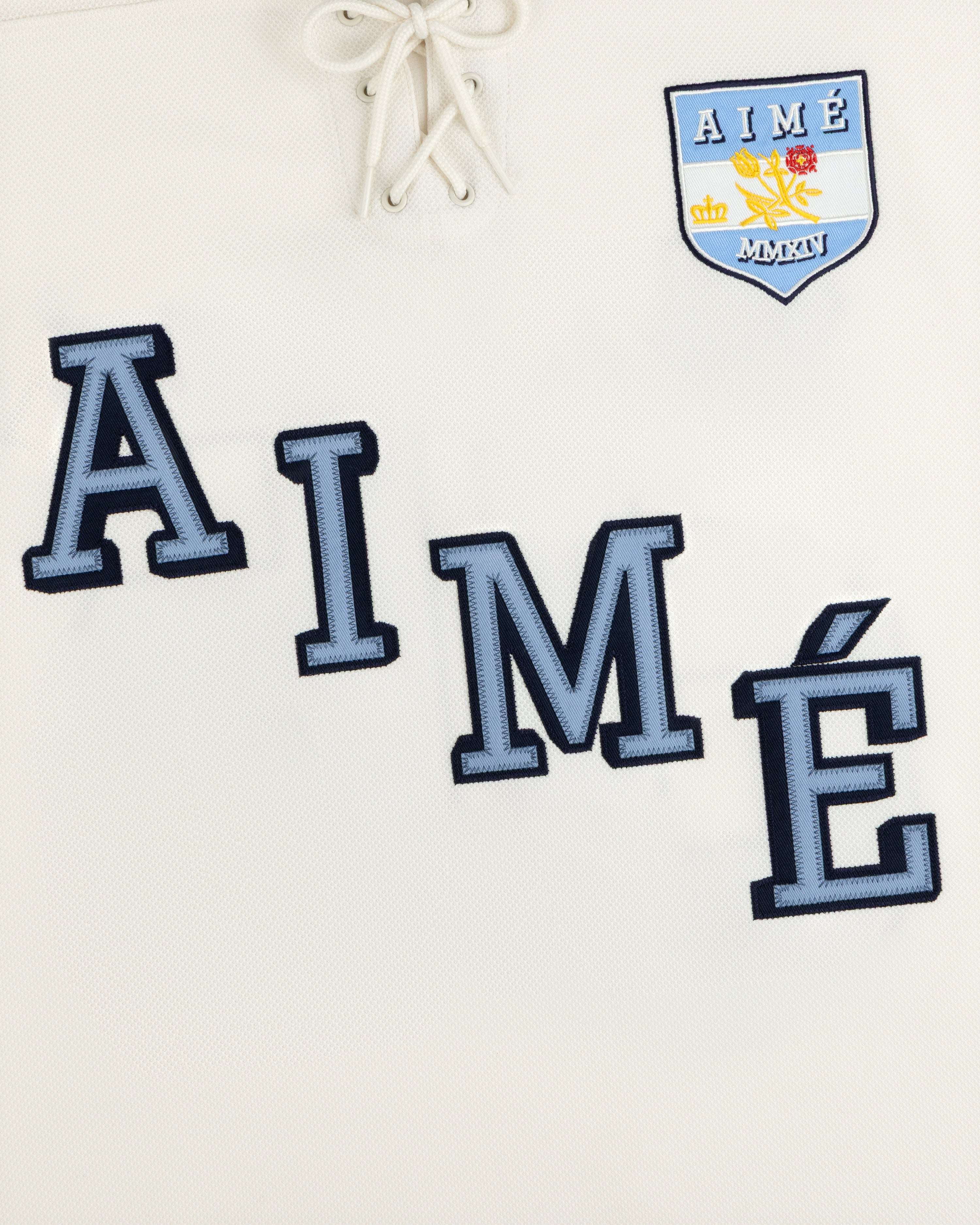Off-white Team Aimé Hockey Jersey with blue AIMÉ lettering, a lace-up collar, and a floral and shield crest.