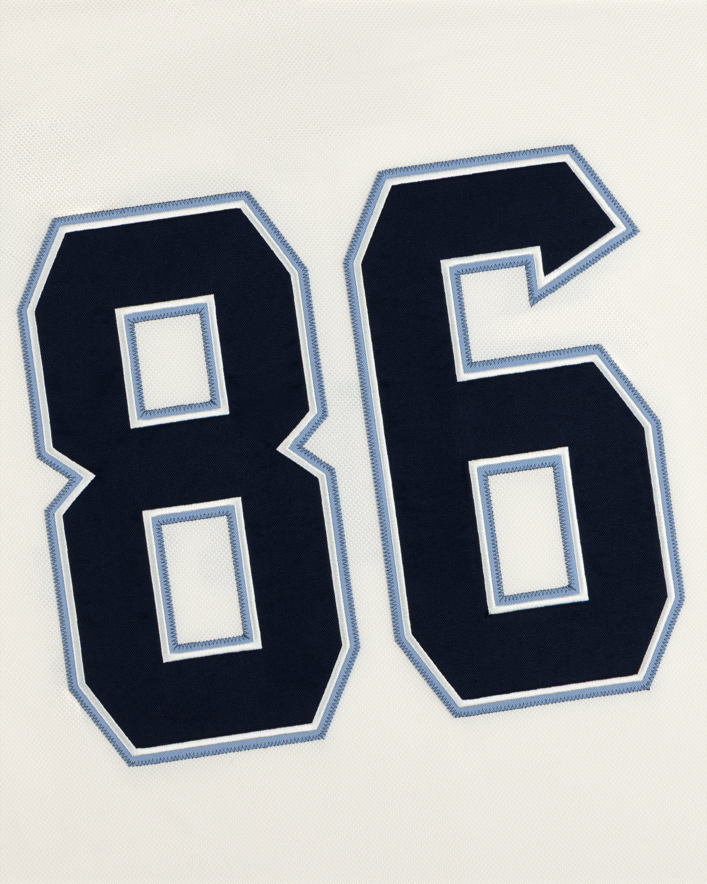 Dark blue athletic number 86 with light blue and white stitched trim on a white Team Aimé Hockey Jersey.