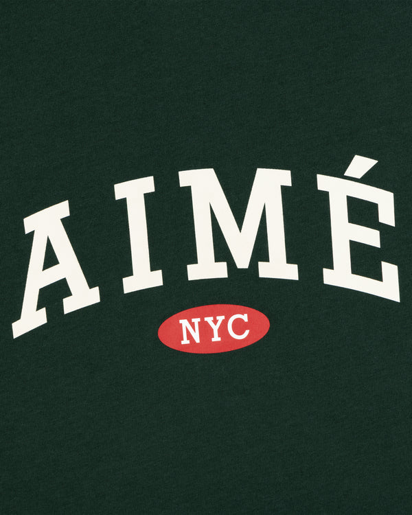 White “AIMÉ” over red oval “NYC” on dark green tee. Aimé Souvenir Tee.