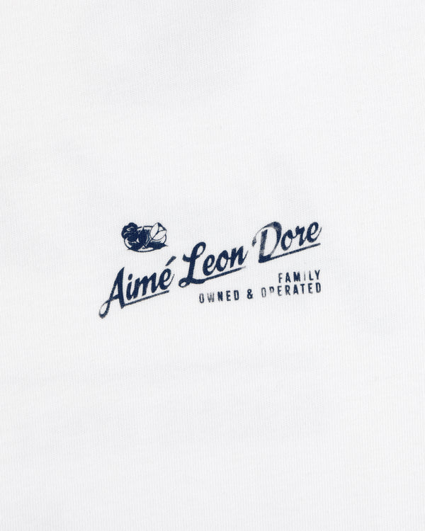 White Neighborhood Tee with a navy Aimé Leon Dore Family Owned & Operated logo and sailing boat graphic above the text.