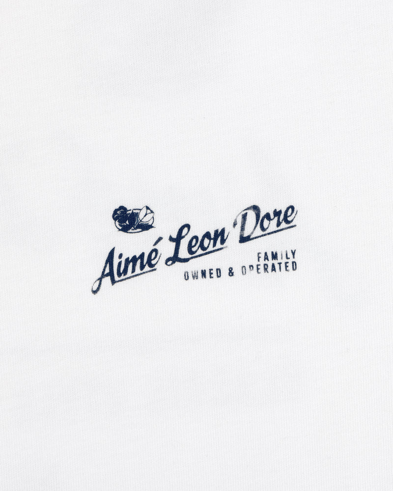 White Neighborhood Tee with a navy Aimé Leon Dore Family Owned & Operated logo and sailing boat graphic above the text.