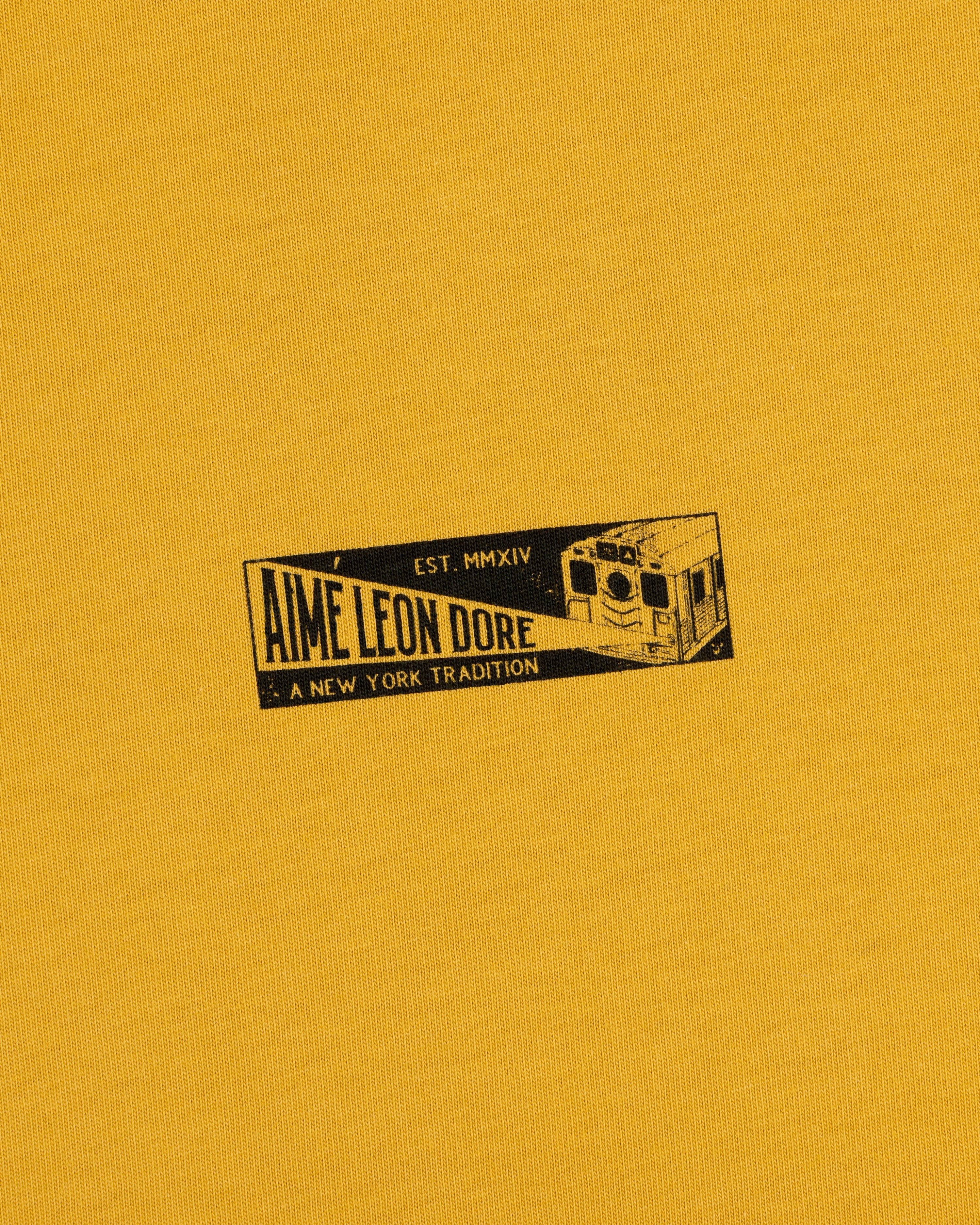 Mustard yellow Aimé Leon Dore Transit Tee with subway car graphic, text: Aime Leon Dore, Est. MMXIV, A New York Tradition.