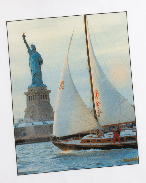 A Long-Sleeve Sailing Tee features a sailboat with red branding on its sail cruising past the Statue of Liberty.