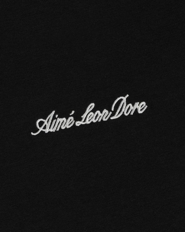 White embroidered Aimé Leon Dore script logo from the Shrunken Script Tee centered on a black fabric background.