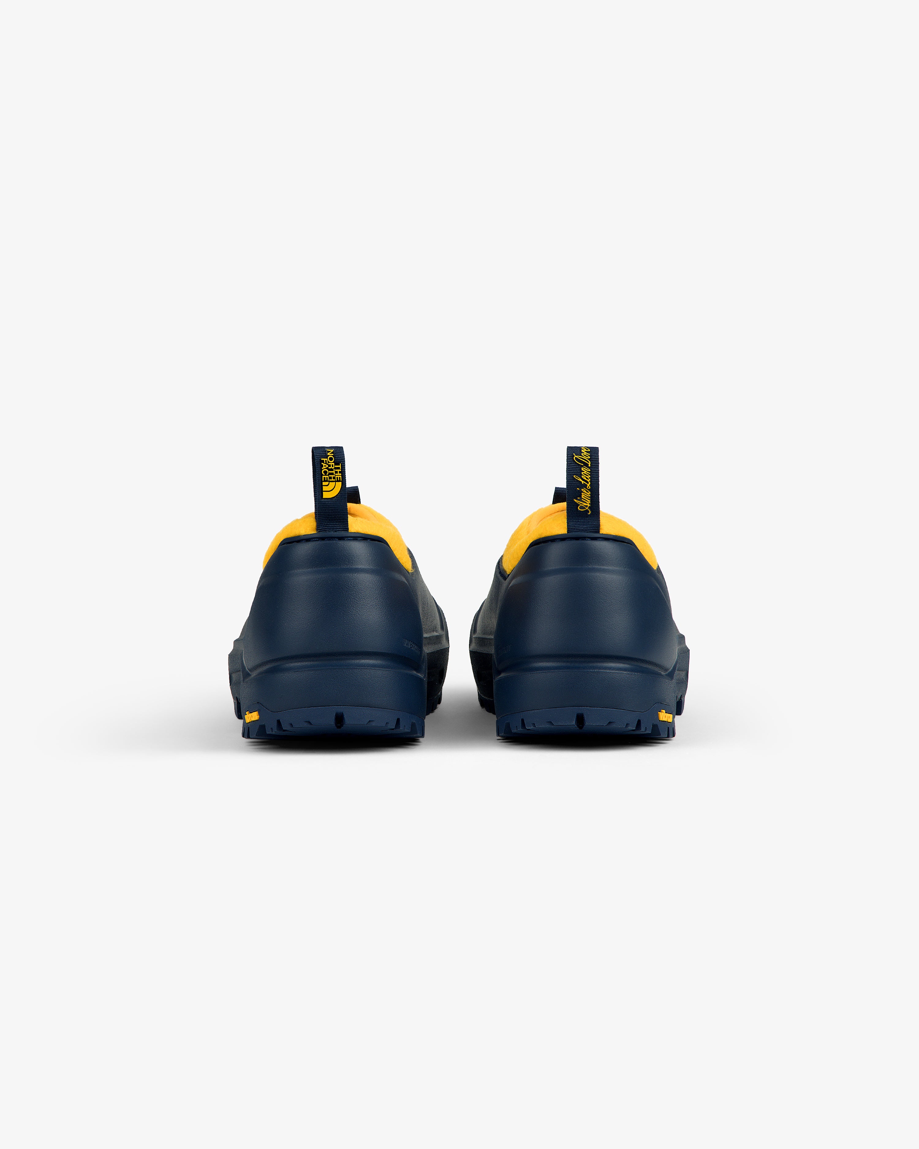 Rear view of ALD / The North Face Base Camp Mule in navy with yellow accents and pull tabs on white background.