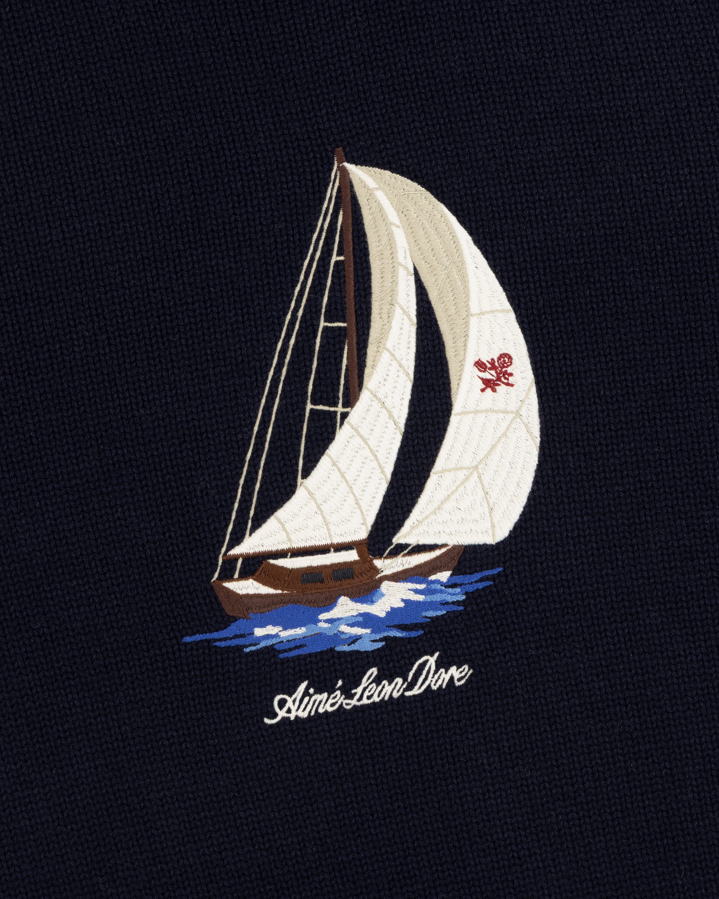 Embroidered sailboat on navy fabric; “Regatta” crewneck sweater by Aimé Leon Dore with cursive branding below.
