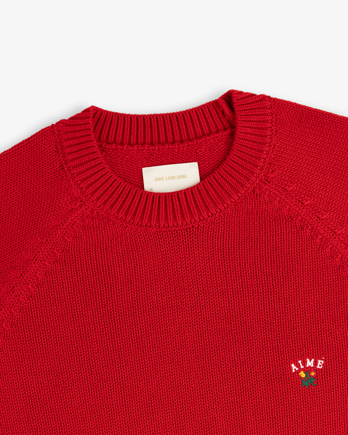Close-up of a red Crest Raglan Sweater with AIME and a small rose embroidered on the chest.