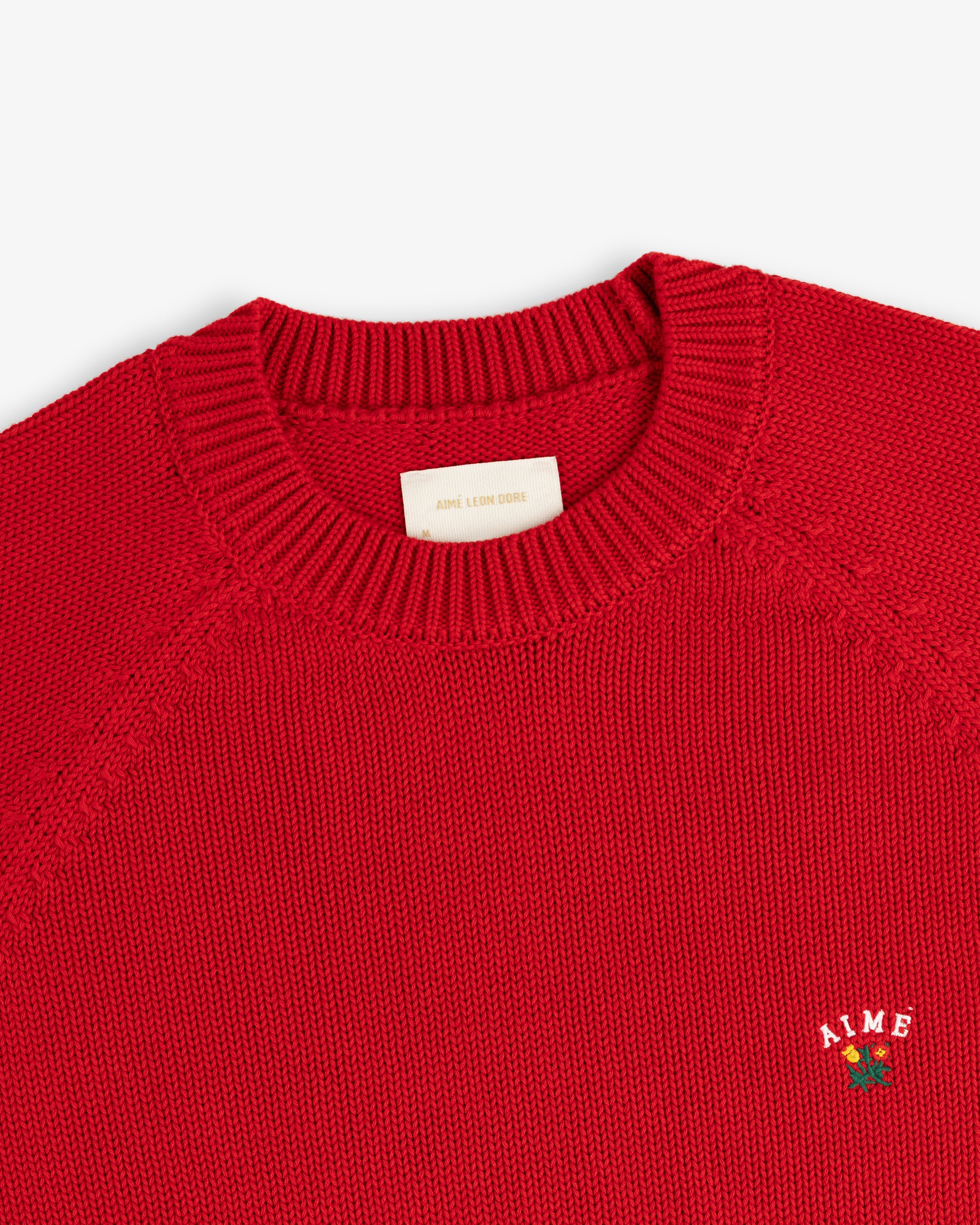Close-up of a red Crest Raglan Sweater with AIME and a small rose embroidered on the chest.