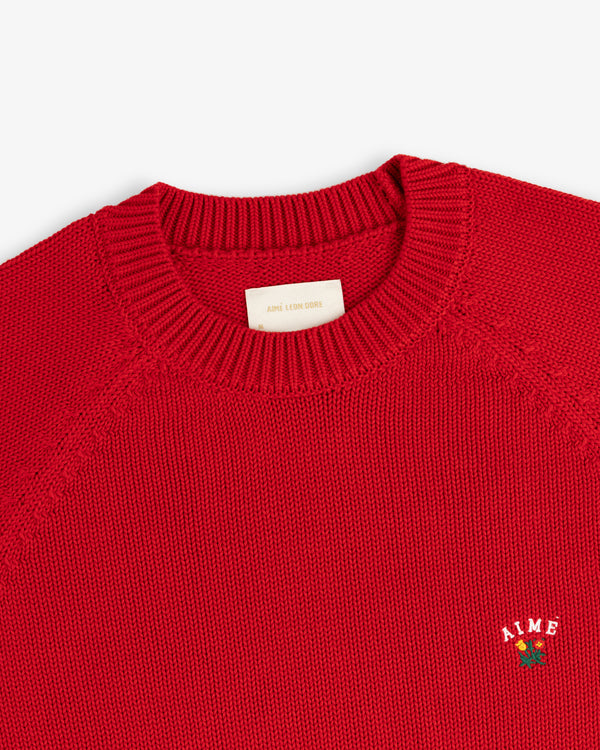 Close-up of a red Crest Raglan Sweater with AIME and a small rose embroidered on the chest.