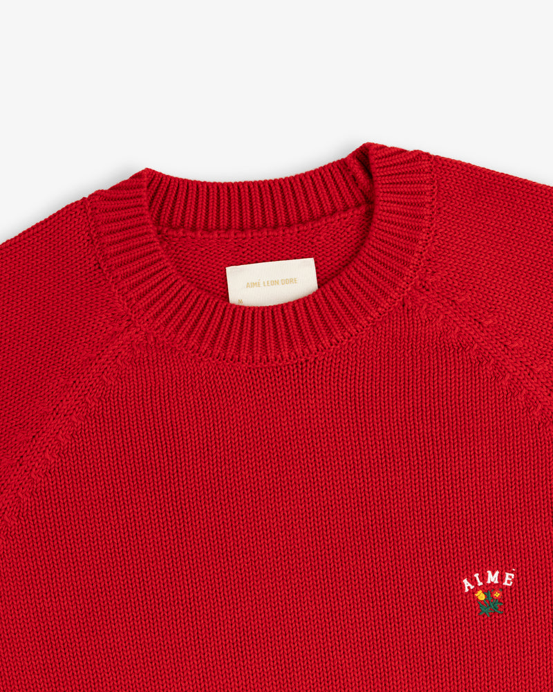 Close-up of a red Crest Raglan Sweater with AIME and a small rose embroidered on the chest.