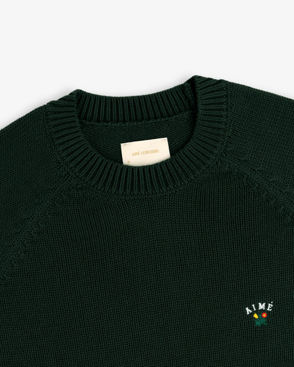 Crest Raglan Sweater in dark green with AIMÉ and rose embroidery on the left chest, shown on white background.