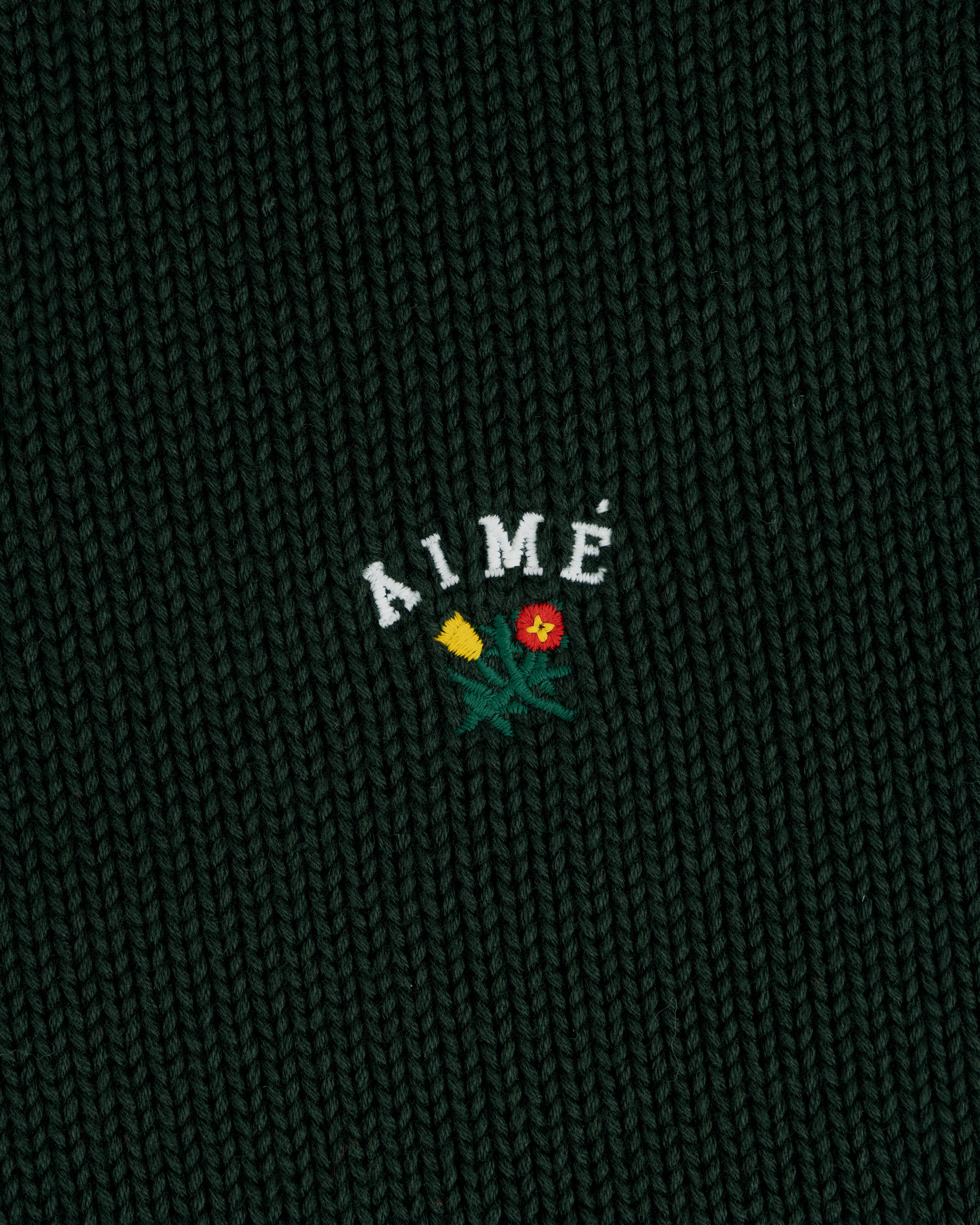 Close-up of dark green Crest Raglan Sweater with AIMÉ and two embroidered flowers, one yellow and one red, below the text.