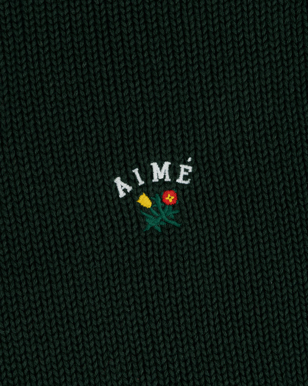 Close-up of dark green Crest Raglan Sweater with AIMÉ and two embroidered flowers, one yellow and one red, below the text.