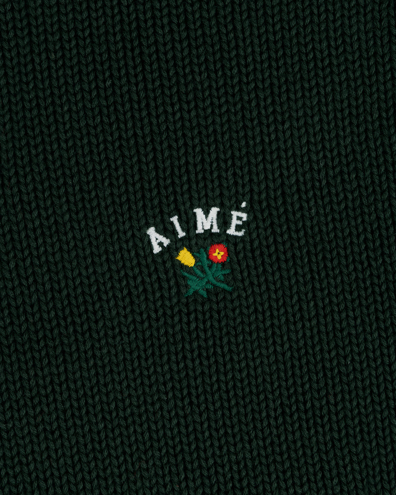 Close-up of dark green Crest Raglan Sweater with AIMÉ and two embroidered flowers, one yellow and one red, below the text.