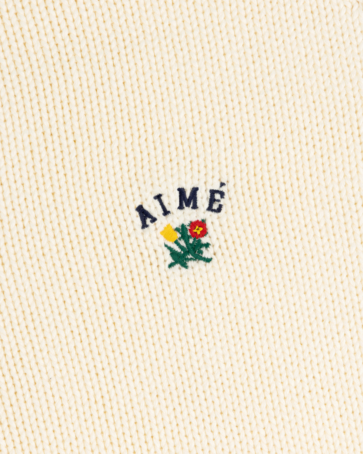 Cream ribbed Crest Raglan Sweater with AIMÉ and a yellow-red flower with green leaves embroidered above.