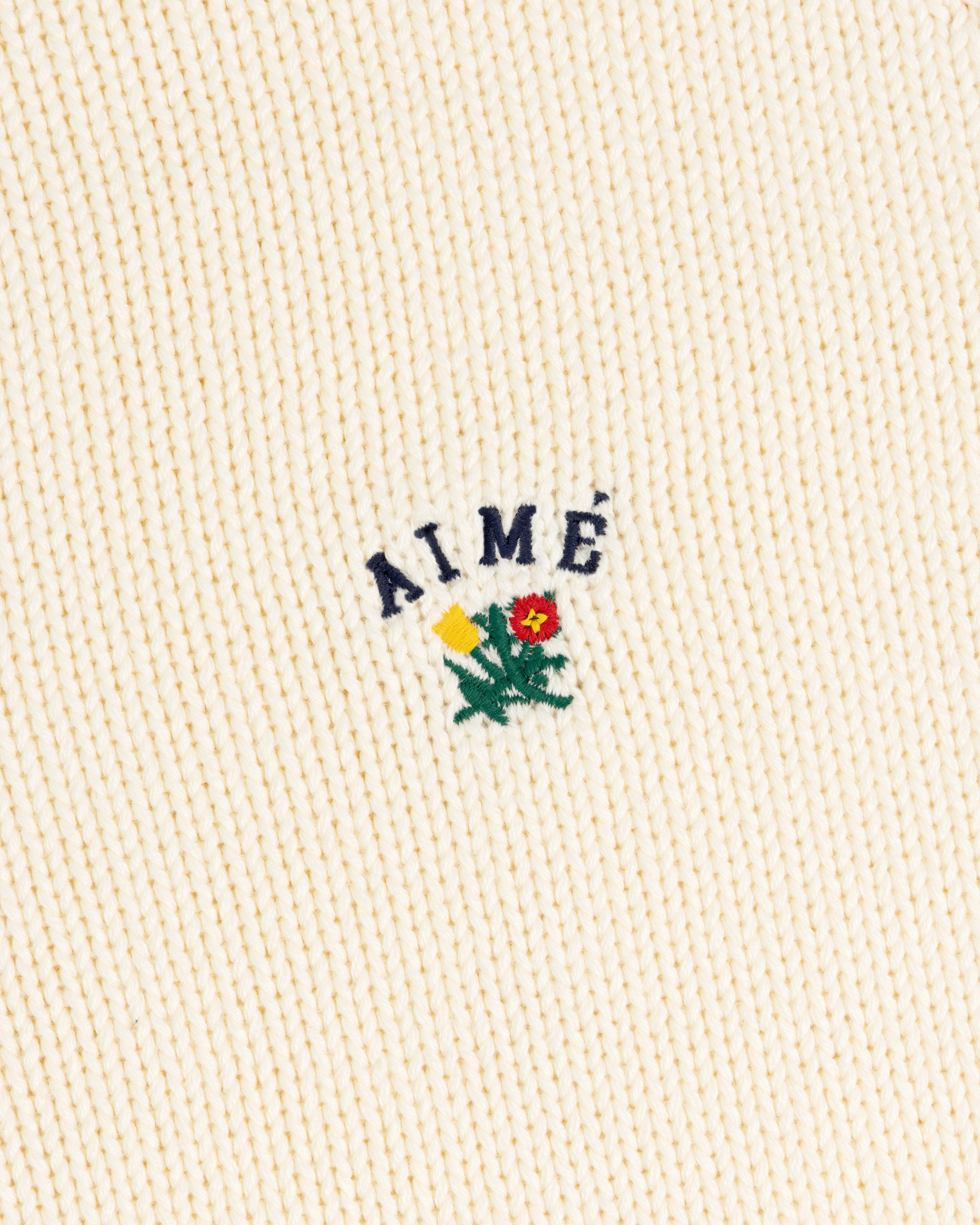 Cream ribbed Crest Raglan Sweater with AIMÉ and a yellow-red flower with green leaves embroidered above.