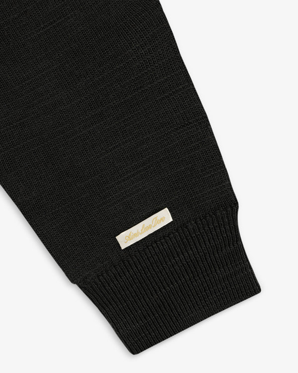 Black Slub Merino Wool Sweater sleeve with a ribbed cuff and a small, rectangular logo tag near the wrist.
