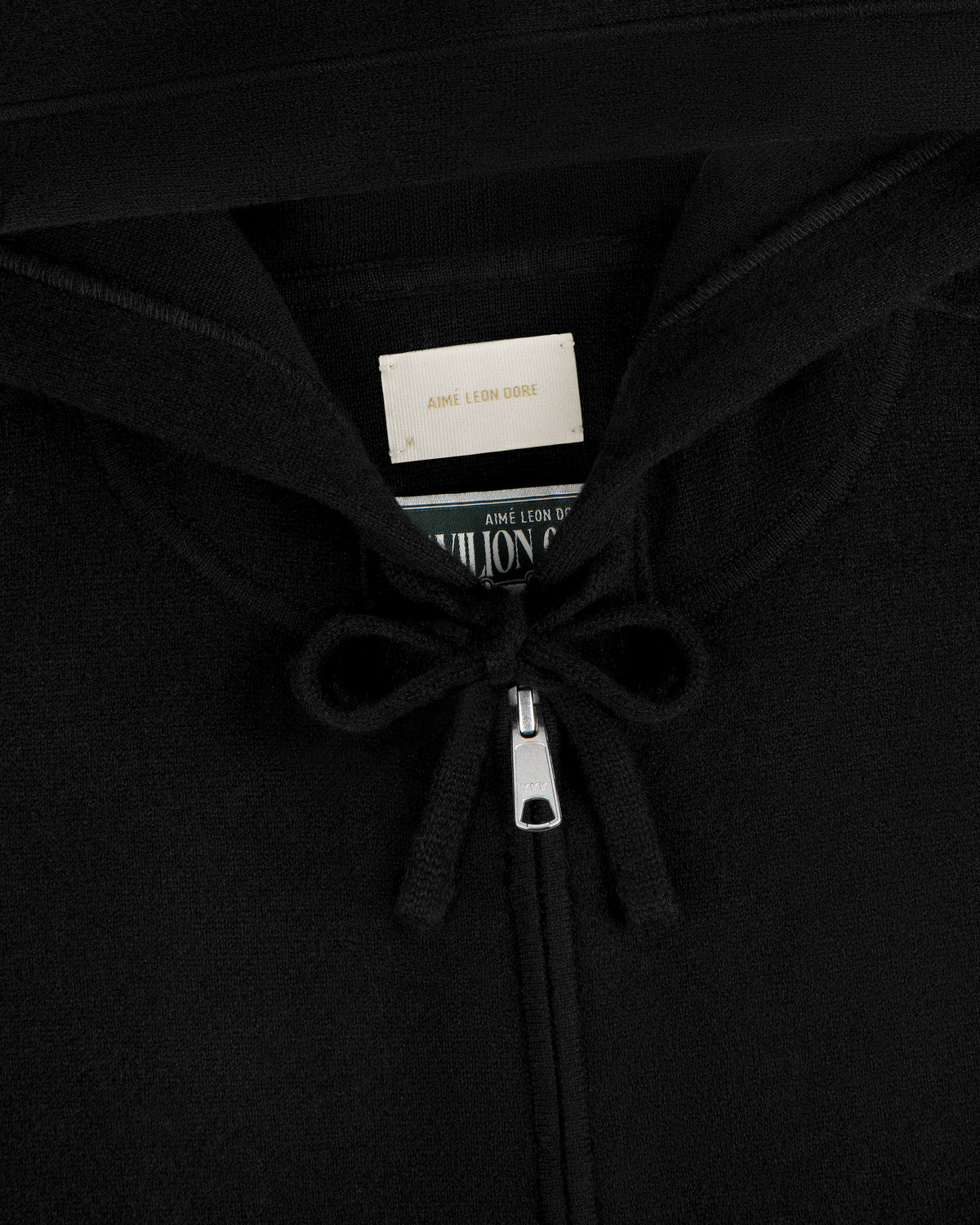 Close-up of the Full-Zip Pavilion Cashmere Hoodie in black, featuring drawstrings and an Aimé Leon Dore label in the collar.