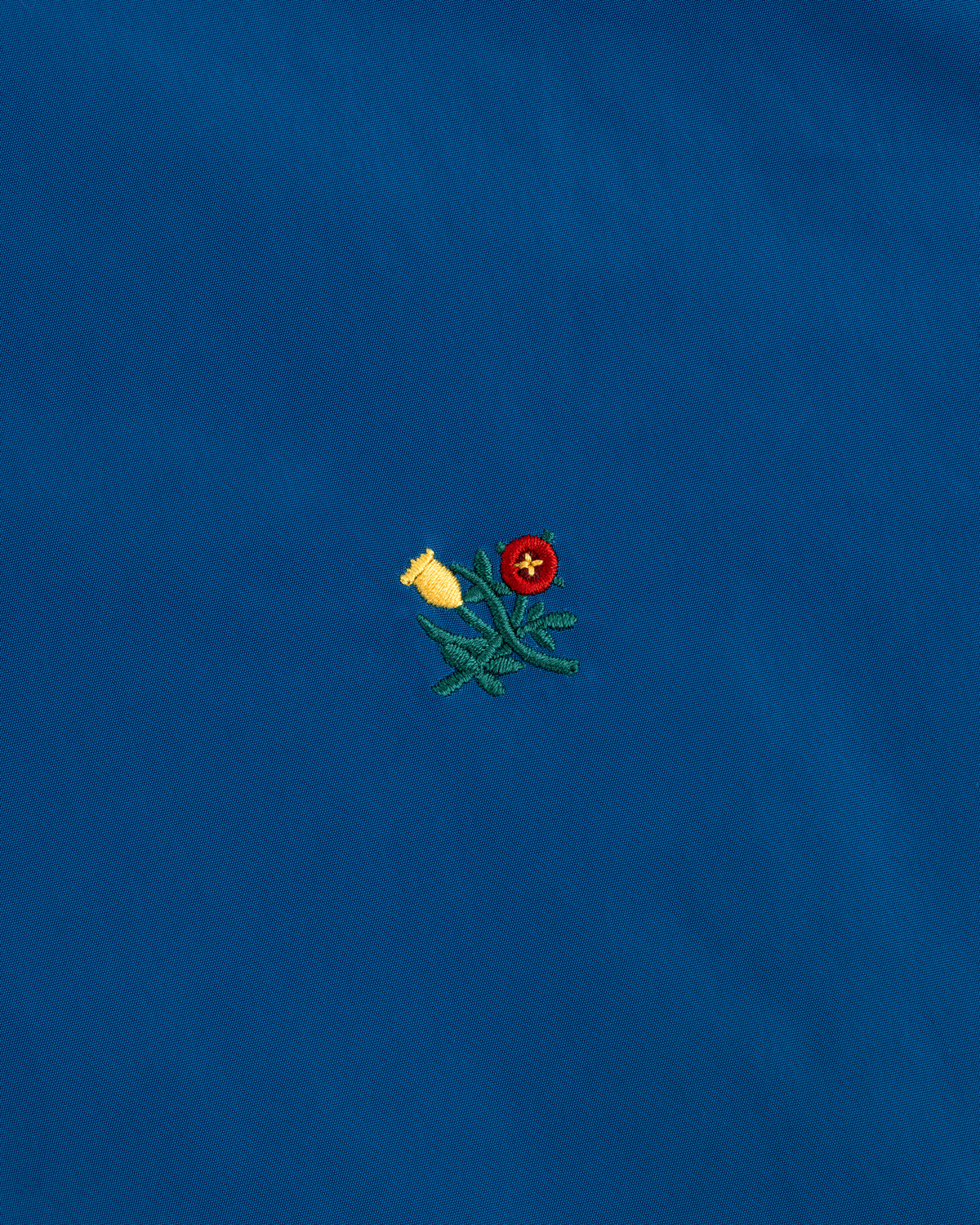 A small yellow and red flower with green leaves, embroidered on a blue Crest Nylon Windbreaker.