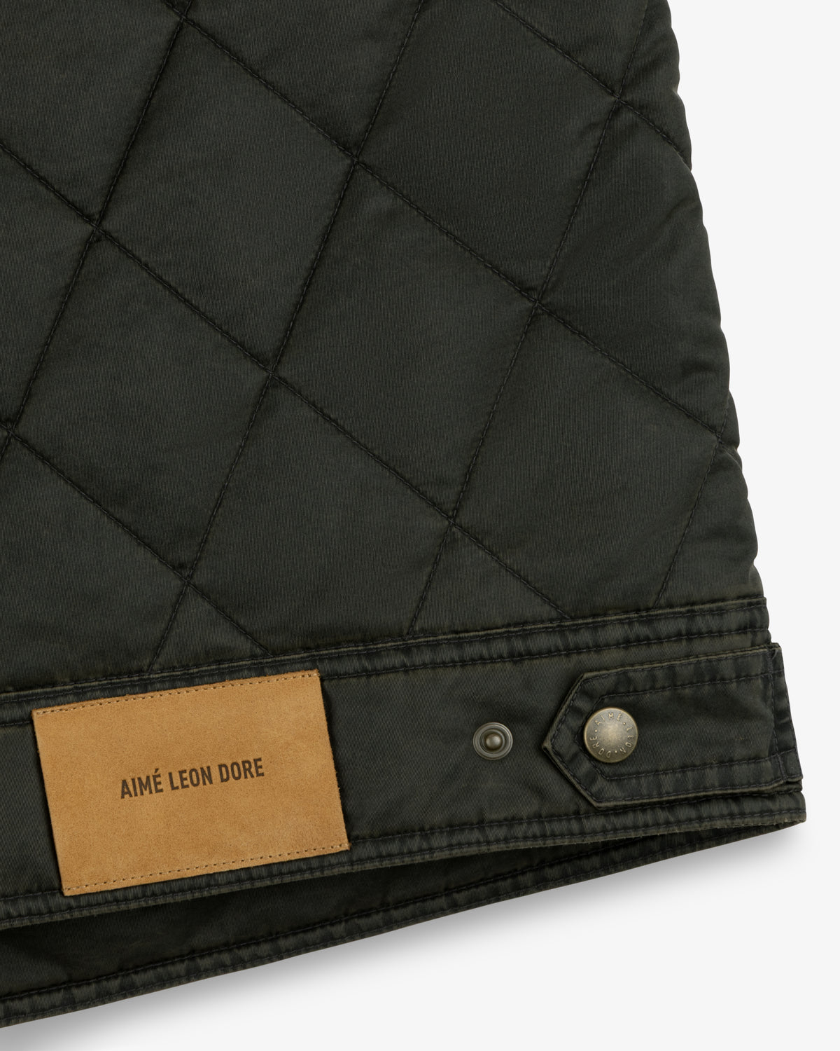 Close-up of a dark green Quilted Trucker Jacket with a tan leather brand patch and a metallic button at the hem.