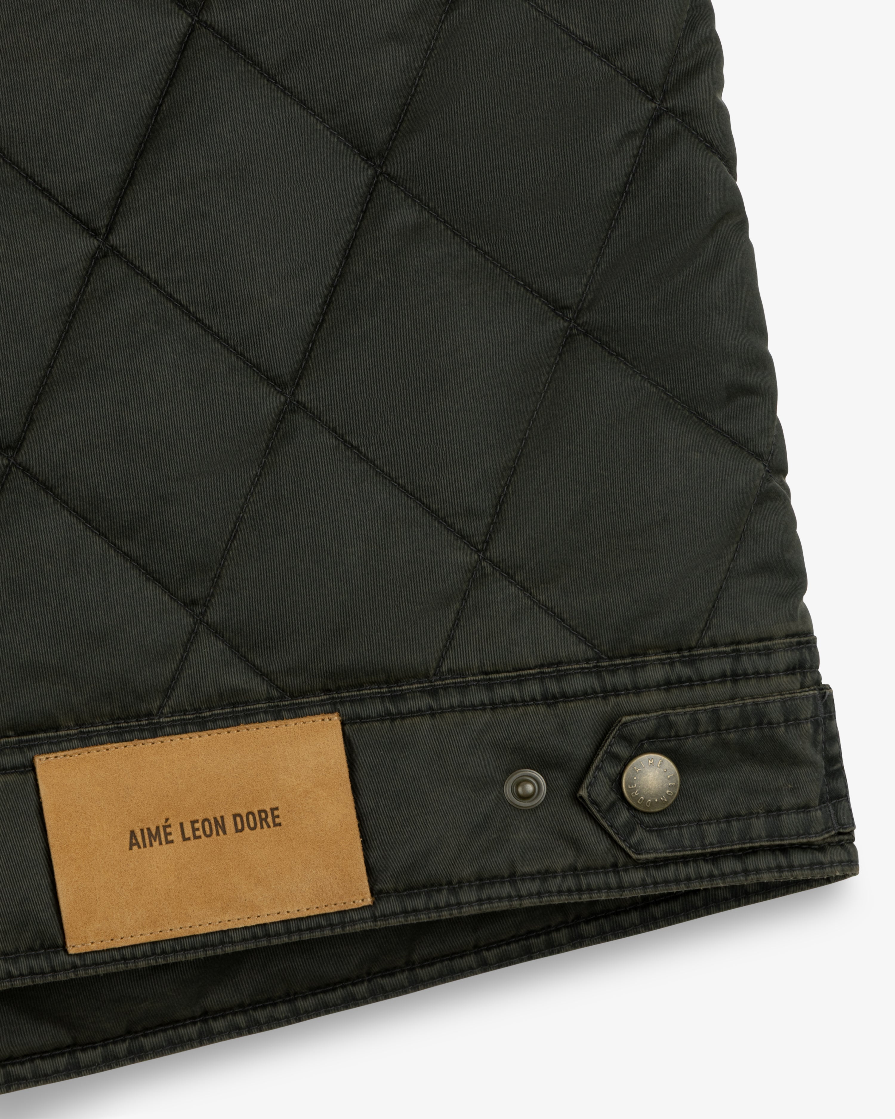 Close-up of a dark green Quilted Trucker Jacket with a tan leather brand patch and a metallic button at the hem.
