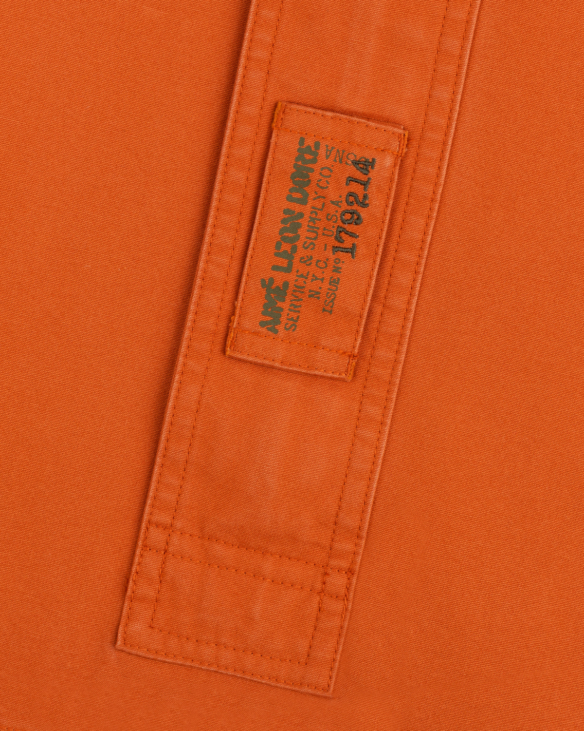 Orange Garment Washed Anorak fabric with a stitched rectangular patch featuring branded text and an issue number.