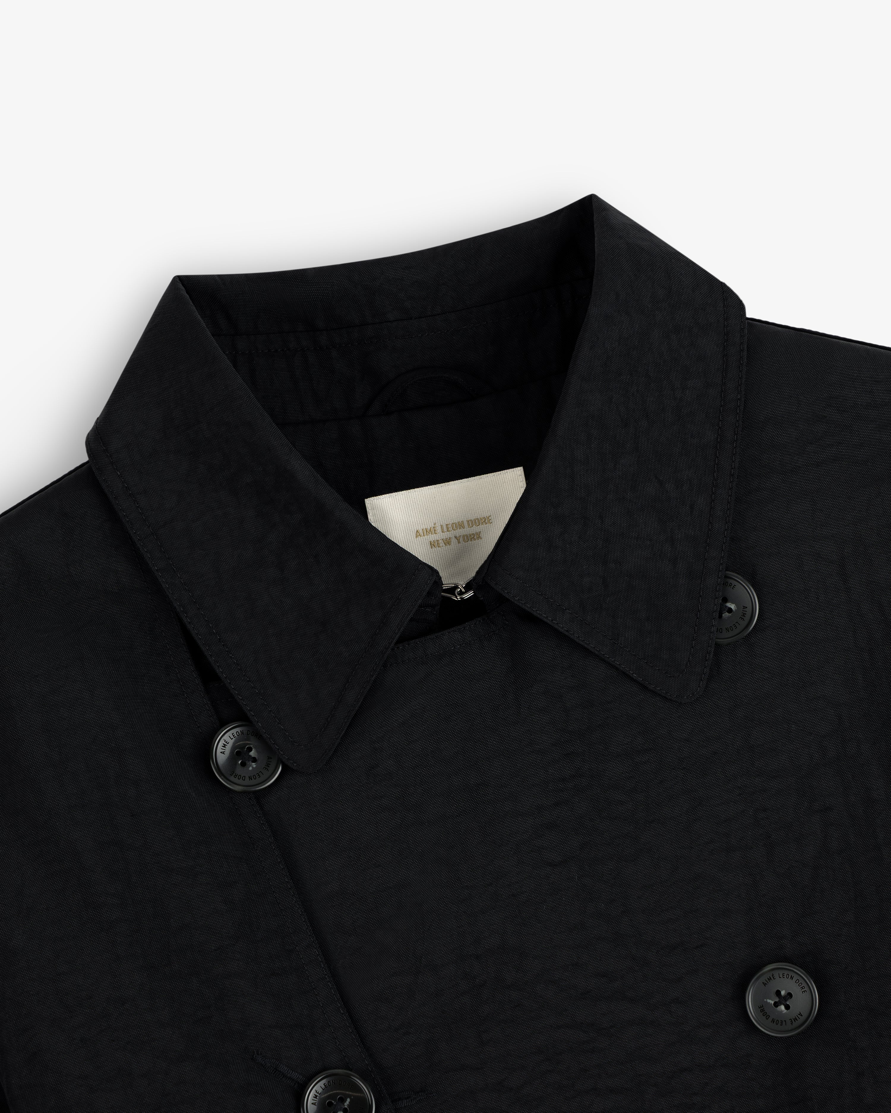 Close-up of a black Double Breasted Nylon Trench Coat with buttons and inner label, on a white background.
