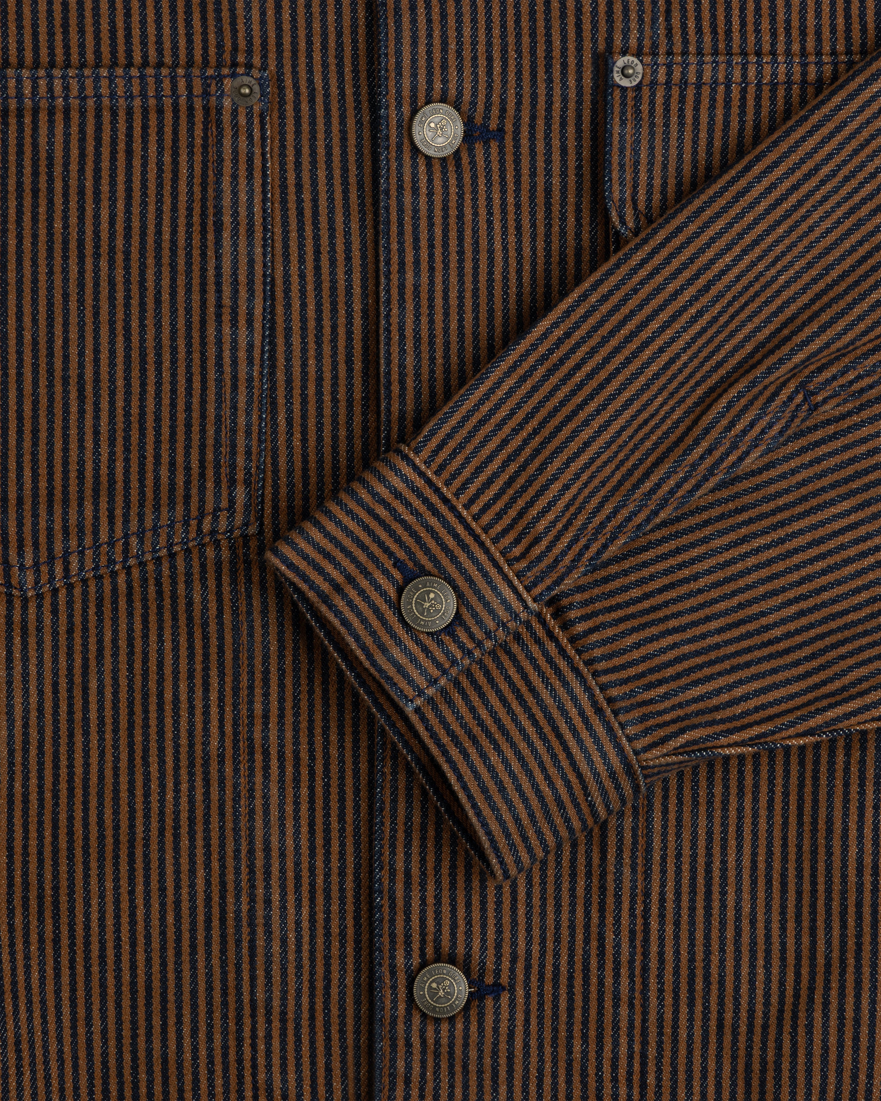 Close-up of a Hickory Stripe Denim Trucker Jacket with brown and navy stripes, metal buttons, pocket, and sleeve.