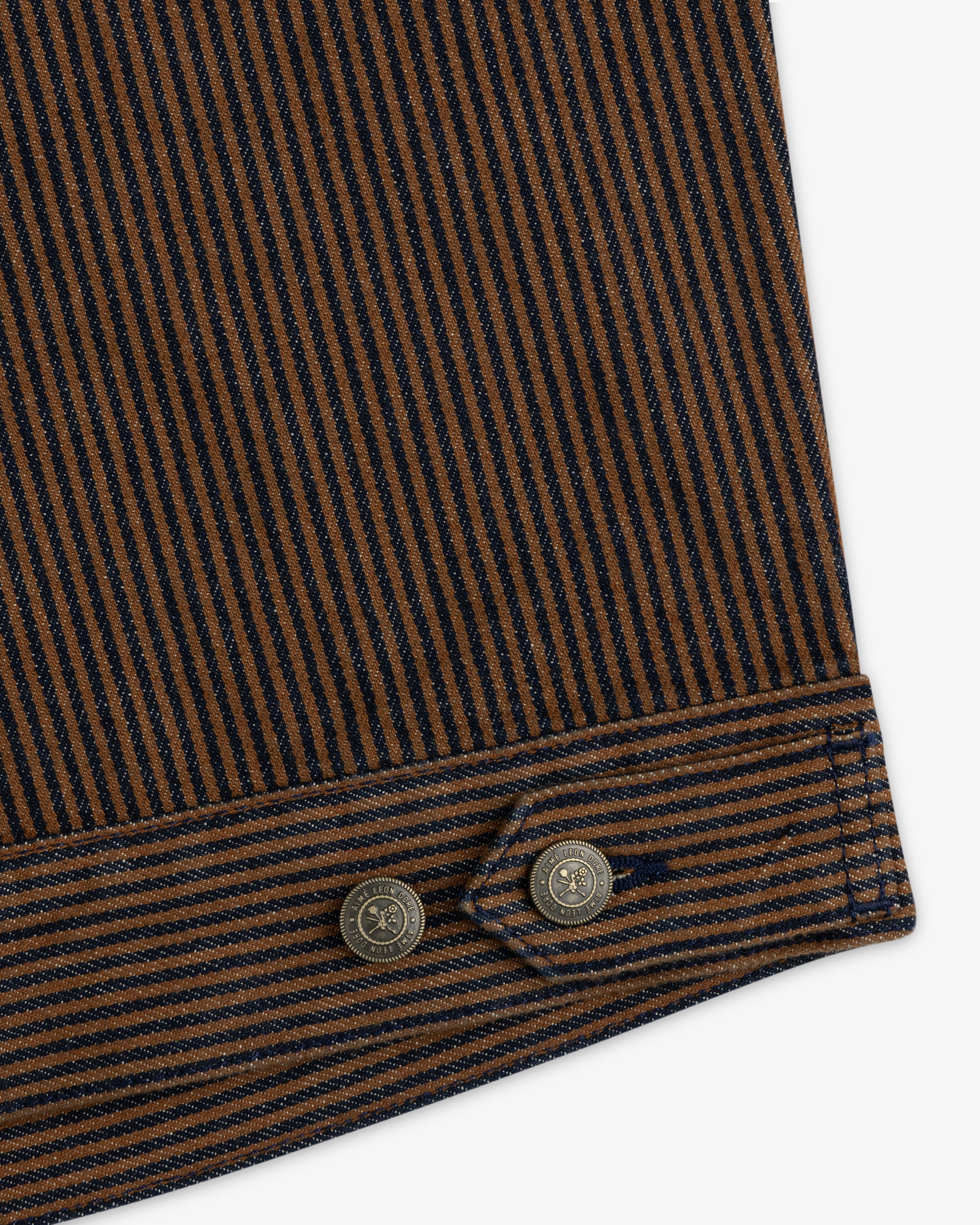 Close-up of the Hickory Stripe Denim Trucker Jacket: brown/navy stripes, stitched edge, and two metal buttons.