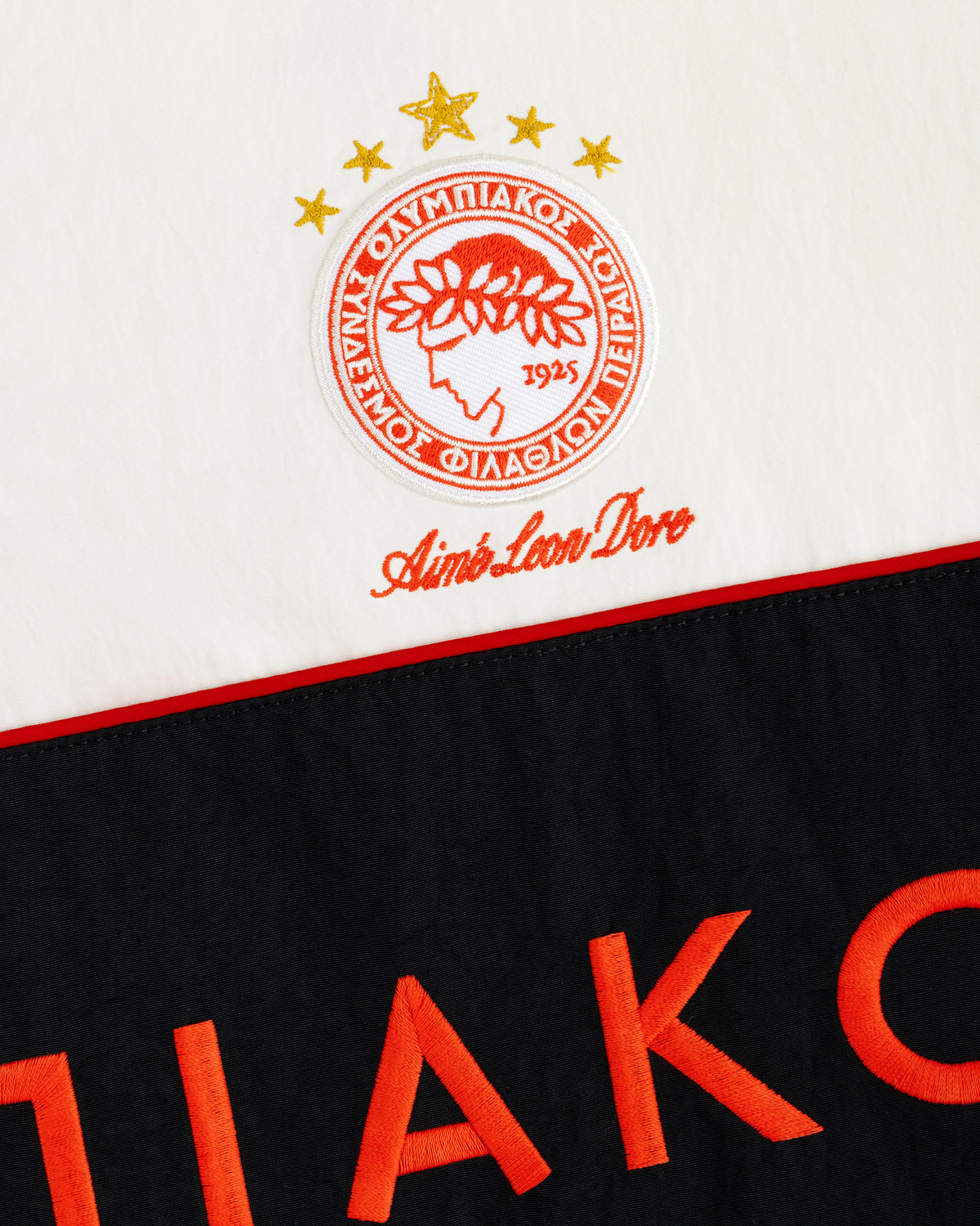 ALD / Olympiacos F.C. Nylon Quarter Zip Pullover in off-white with an embroidered crest and orange lettering on black.