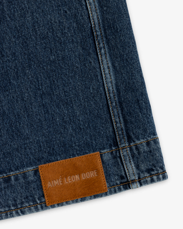 Dark blue denim fabric of a Denim Chore Coat with a tan leather patch embossed with Aimé Leon Dore.