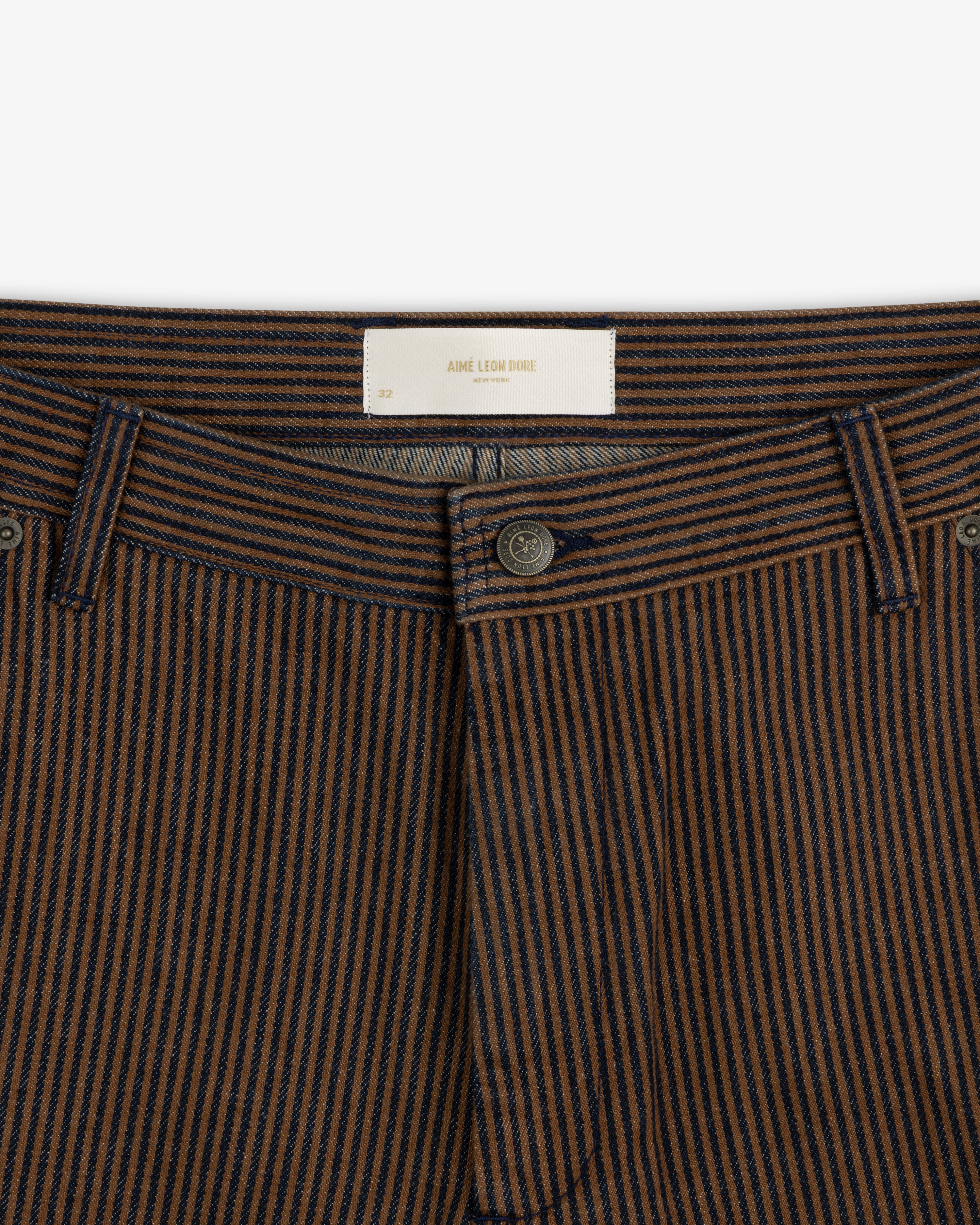Close-up of Hickory Stripe Painter Pant featuring brown and navy stripes, button fly, and white Aime Leon Dore label.