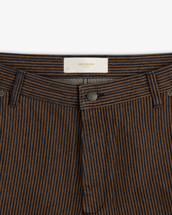 Close-up of Hickory Stripe Painter Pant featuring brown and navy stripes, button fly, and white Aime Leon Dore label.