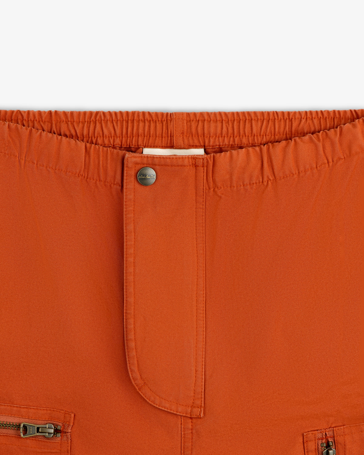 Close-up of rust-colored Garment Washed Aviator Pants with an elastic waist, front button, and zippered pockets.