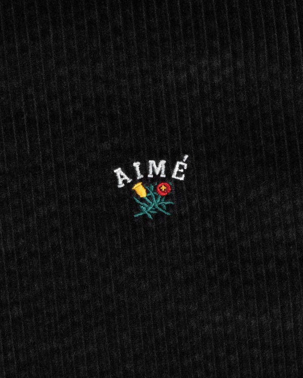 Black corduroy fabric of the Corduroy Leon Overshirt with an embroidered logo featuring AIMÉ above a small flower.